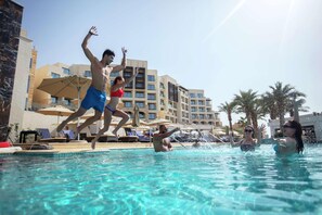 A swimming pool with people jumping, lounge chairs, umbrellas, and a hotel building in the background.