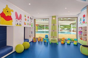Une salle de jeux pour enfants avec des jouets colorés, des sièges et une vue sur l’espace extérieur de la piscine.