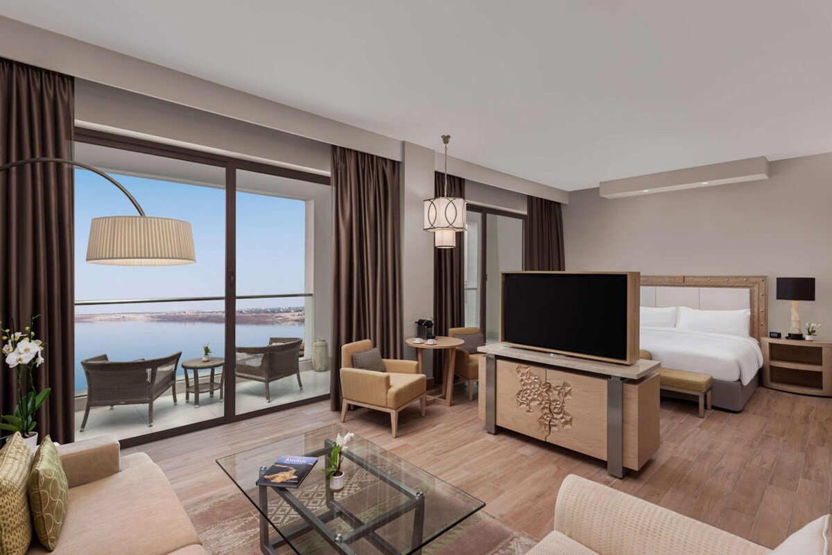 A hotel room with a large bed, a TV, a sofa, a coffee table, and a view of the water.