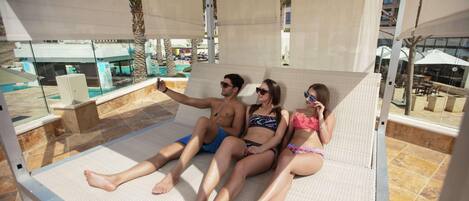 A couple and a child relaxing on a sunbed with a pool view.