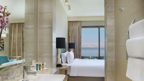 A hotel room with a large bed, a bathroom with a sink and mirror, and a view of the mountains. - Hilton Dead Sea Resort & Spa (Sweimeh)
