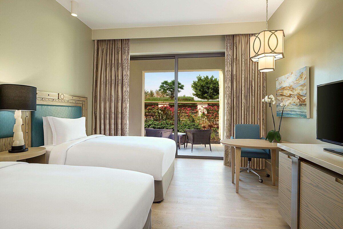 A hotel room with two beds, a desk, a TV, and a balcony with a view of greenery and a building.