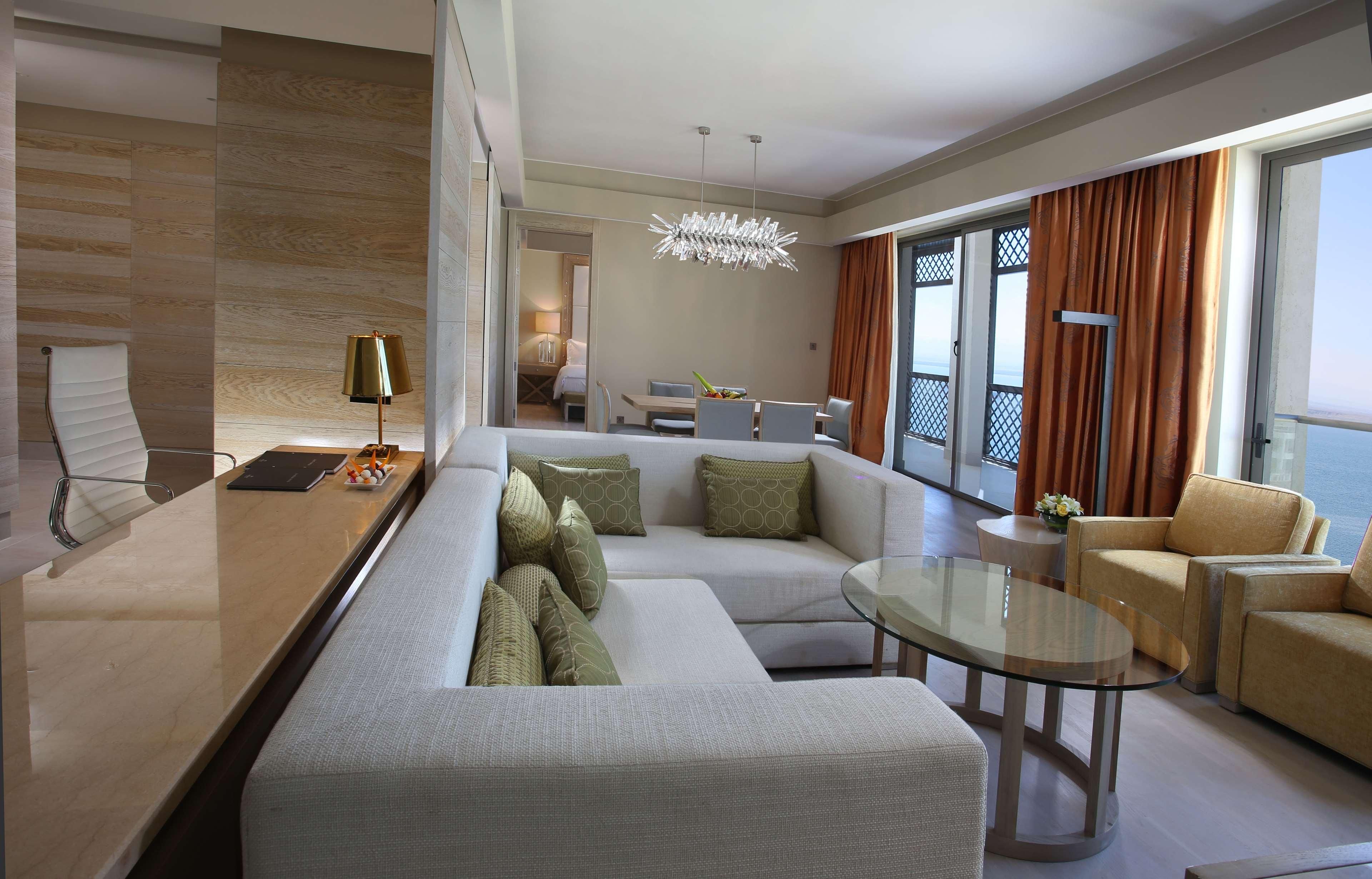 Et moderne hotelværelse med sofa, lænestol, sofabord i glas og et spiseområde med bord og stole.