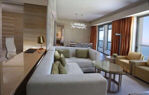 A modern hotel room with a sofa, armchair, glass coffee table, and a dining area with a table and chairs. - Hilton Dead Sea Resort & Spa (Sweimeh)