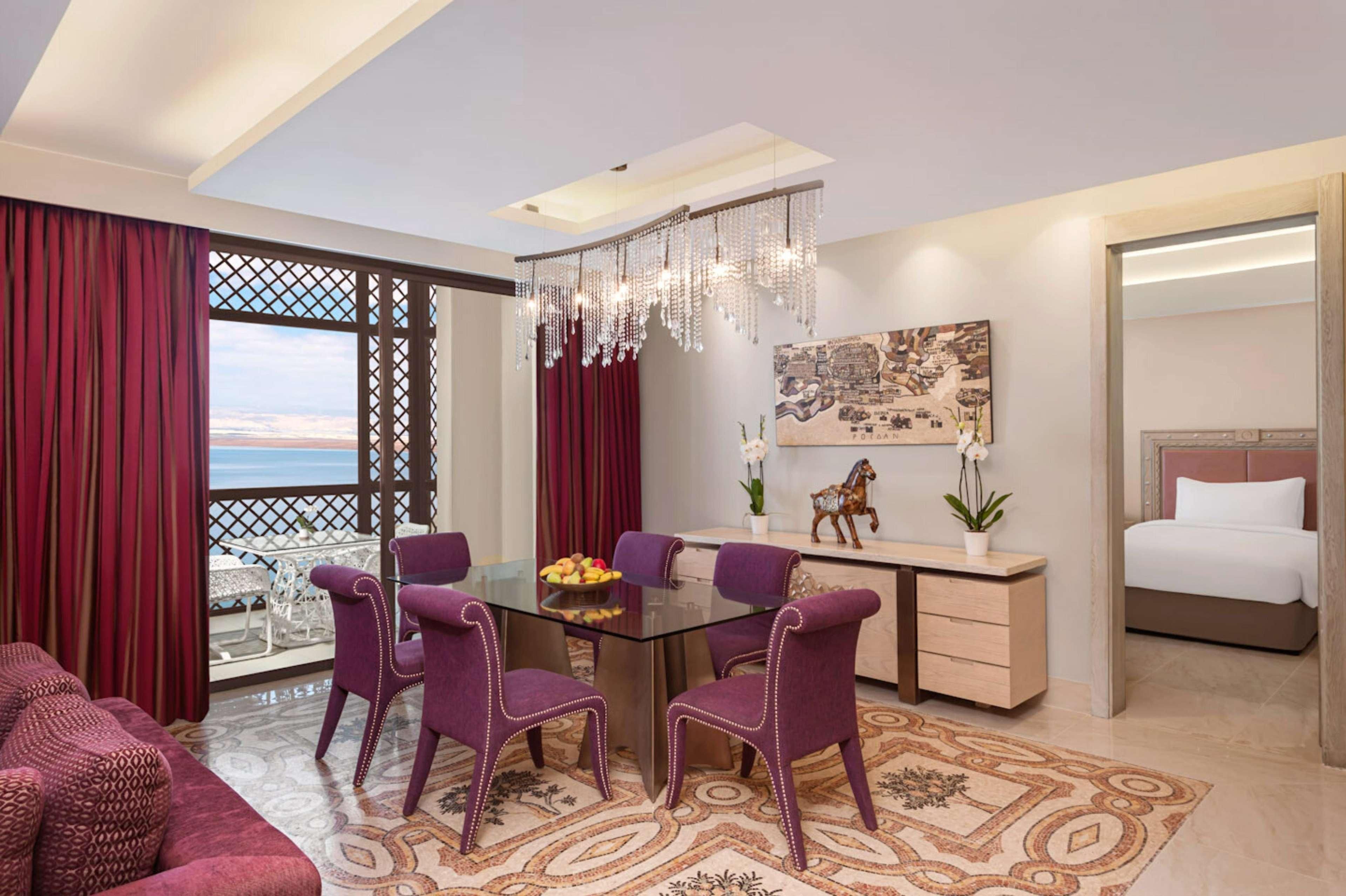 A modern hotel room with a balcony, a dining area with purple chairs, a bed, and a decorative wall piece.