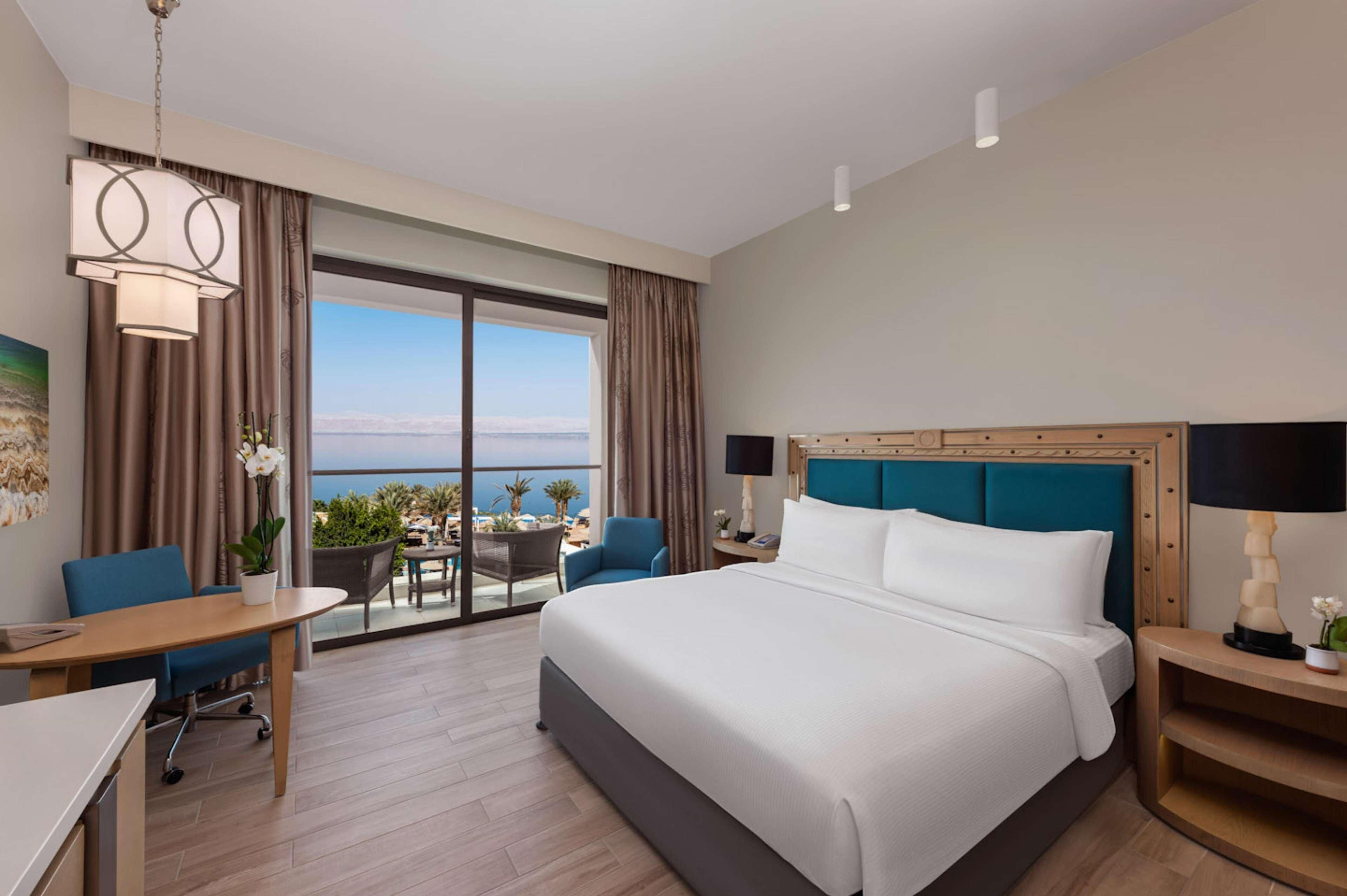 A hotel room with a large bed, a desk, a chair, a table, a lamp, and a view of the ocean.