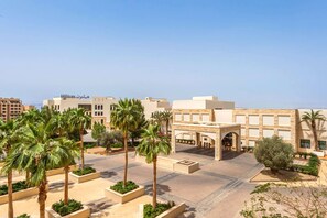 A hotel with a modern architectural design, featuring a central entrance with a covered walkway, palm trees, and a clear sky. - Hilton Dead Sea Resort & Spa (Sweimeh)