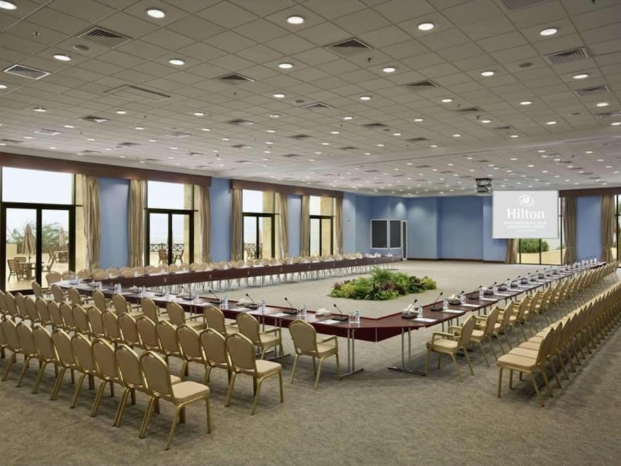 Un amplio salón de conferencias con una larga mesa, filas de sillas y una pantalla para proyector.
