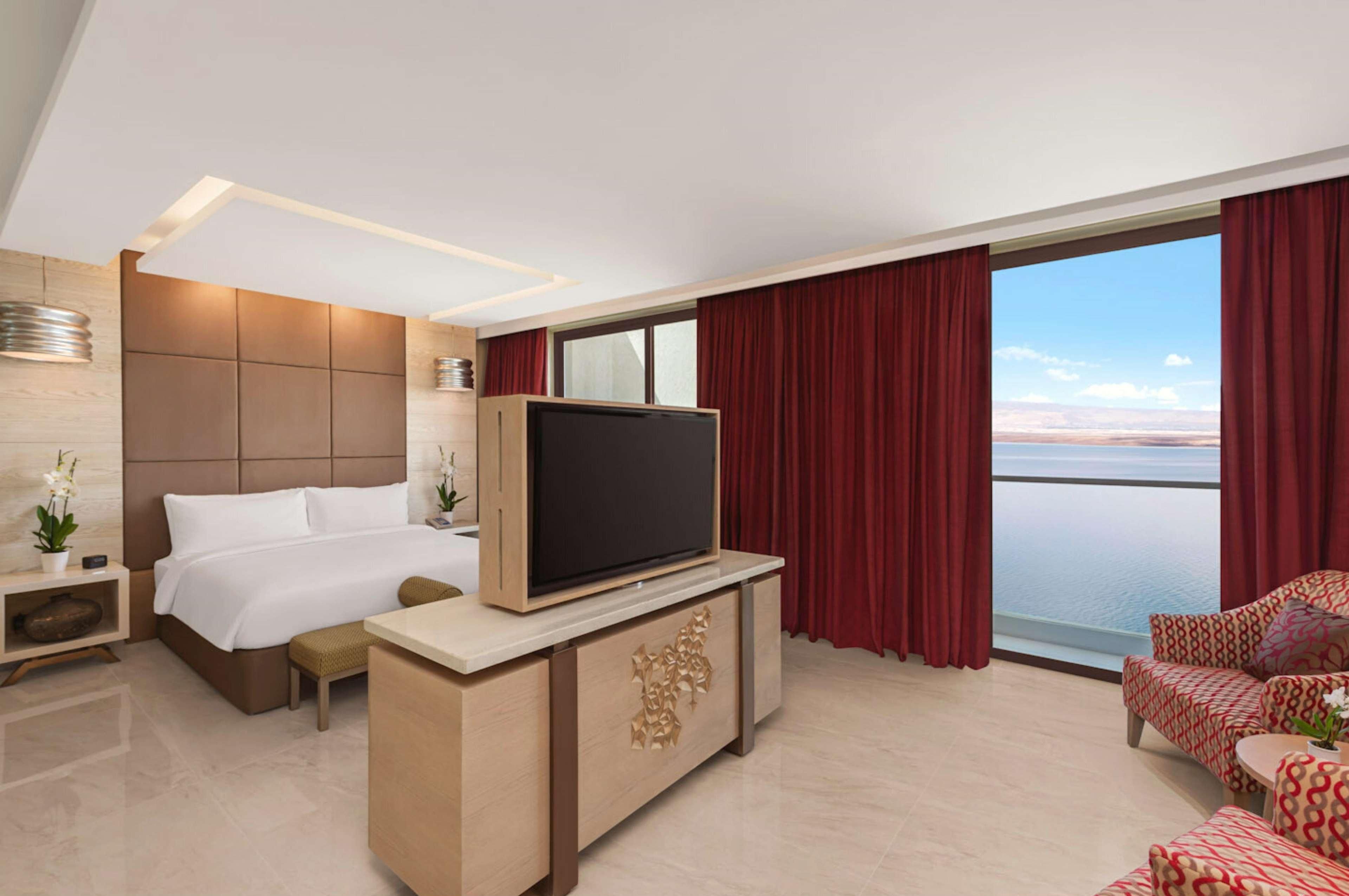A hotel room with a bed, a TV on a wooden stand, a seating area with patterned furniture, and a large window with curtains.