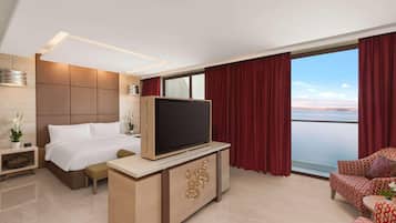 A hotel room with a bed, a TV on a wooden stand, a seating area with patterned furniture, and a large window with curtains.