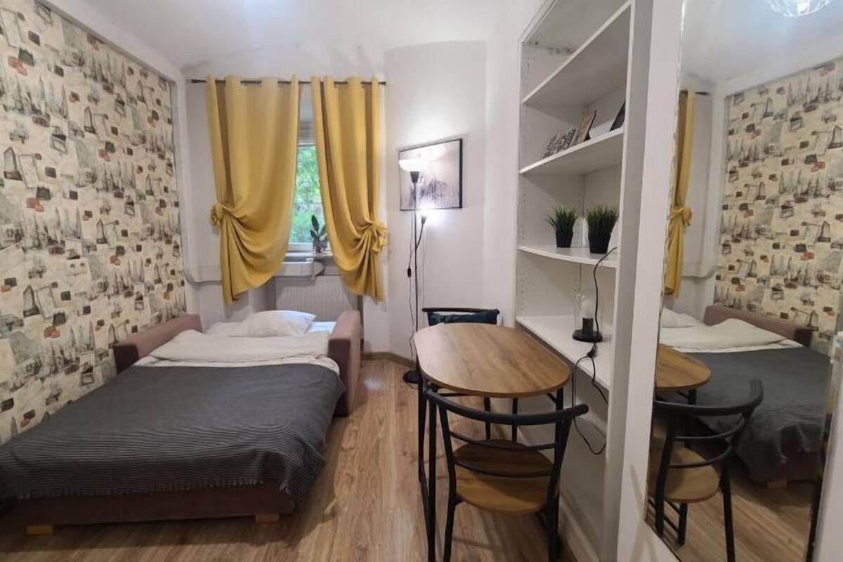 A small, compact room with two single beds, a wooden table with chairs, a shelf with plants, and a window with yellow curtains.