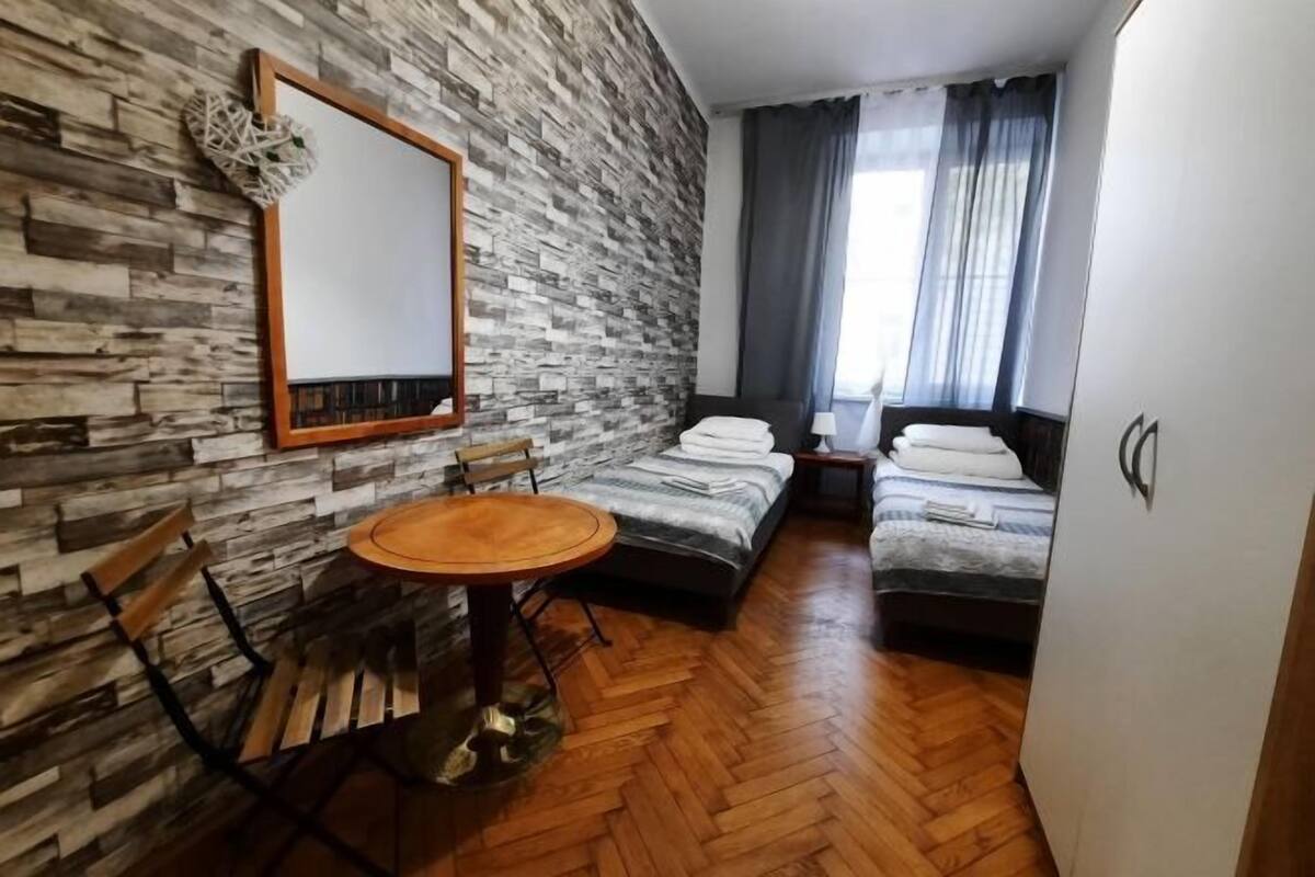 A room with a herringbone-patterned stone wall, a round wooden table with chairs, two single beds, and a large mirror.