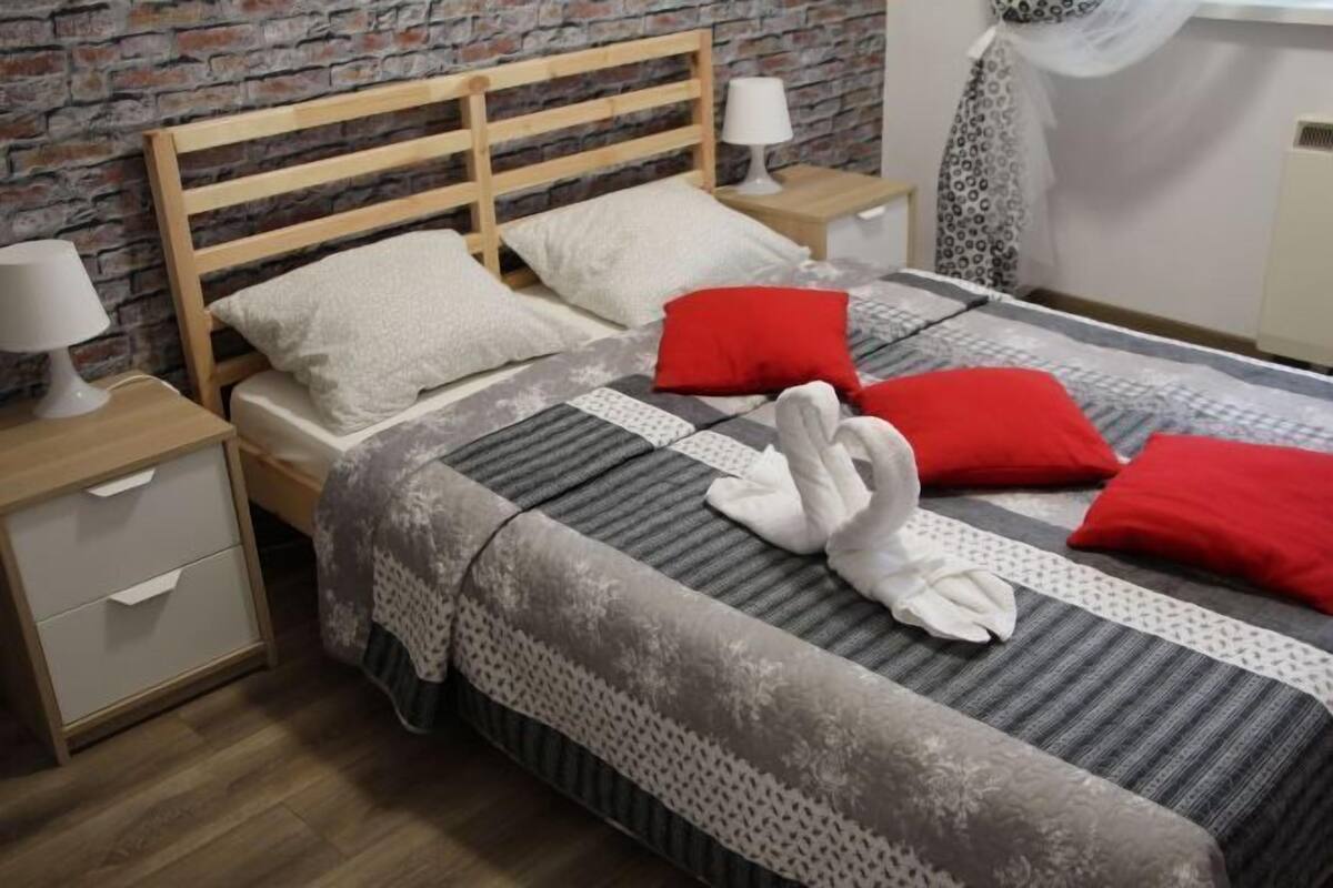 A neatly made bed with a patterned bedspread, two pillows, and a red accent pillow. There is a wooden nightstand with a lamp and a drawer. The room has a brick wall and a window with a white curtain.