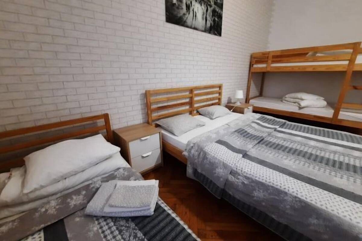A room with a white brick wall, a wooden bed with a headboard, a nightstand, a wooden bunk bed, and a painting on the wall.