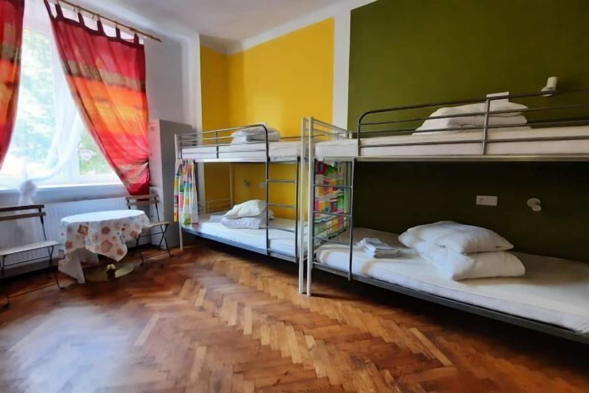 A dormitory room with bunk beds, a small table with a floral tablecloth, and a window with red curtains.
