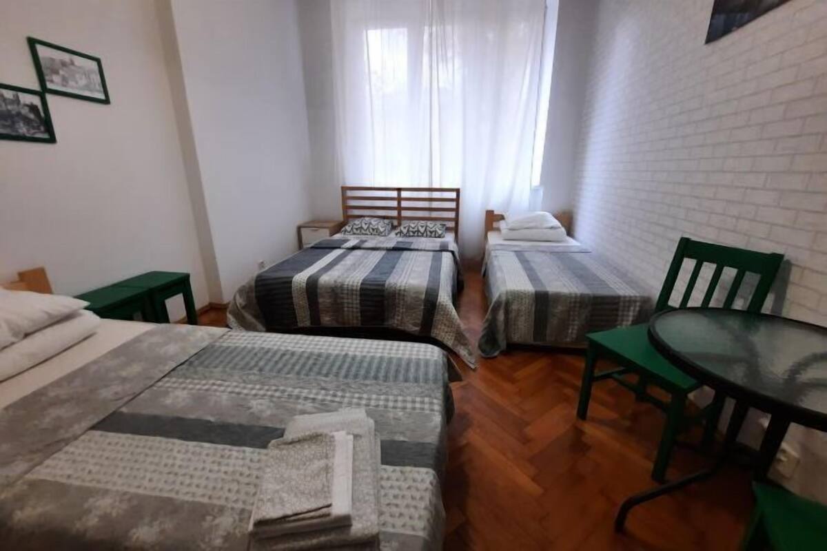 A room with two beds, a small table, a chair, and a window with sheer curtains.