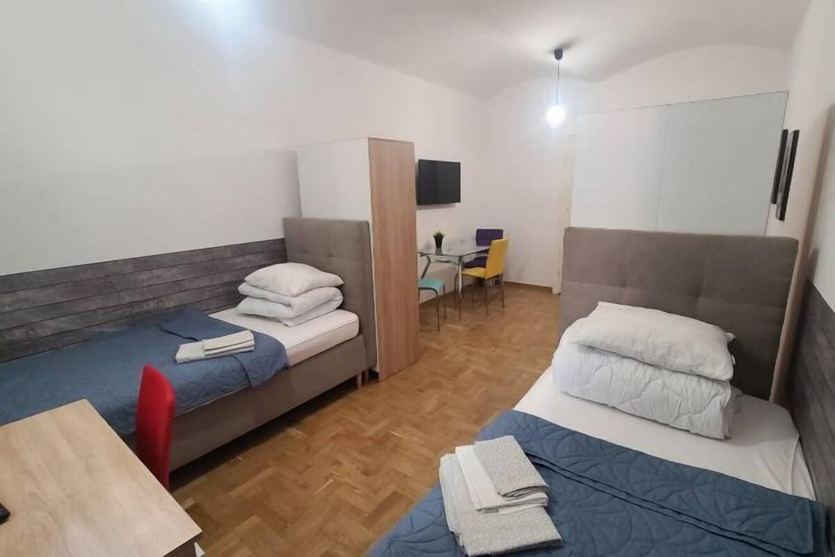A compact hotel room with two single beds, a small table, a chair, and a TV mounted on the wall.