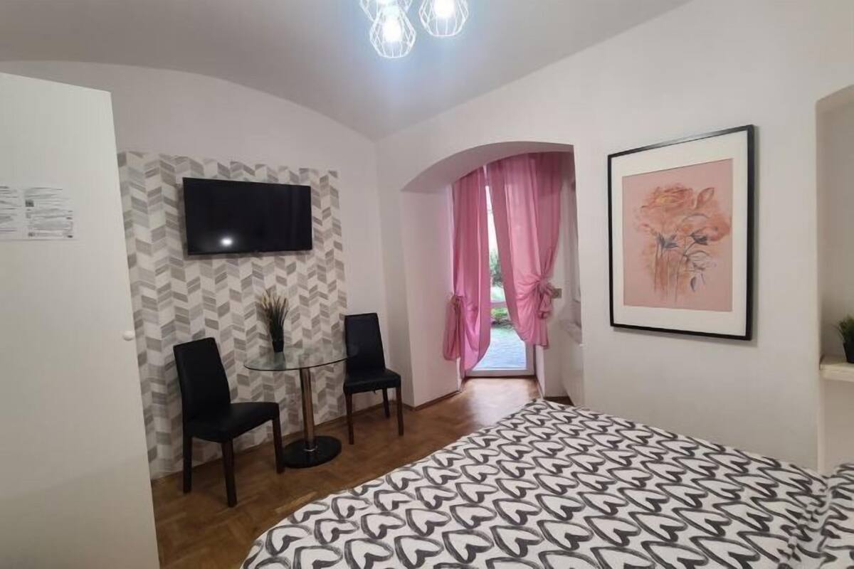 A bedroom with a bed, a TV, a desk with two chairs, a framed picture on the wall, and a window with pink curtains.