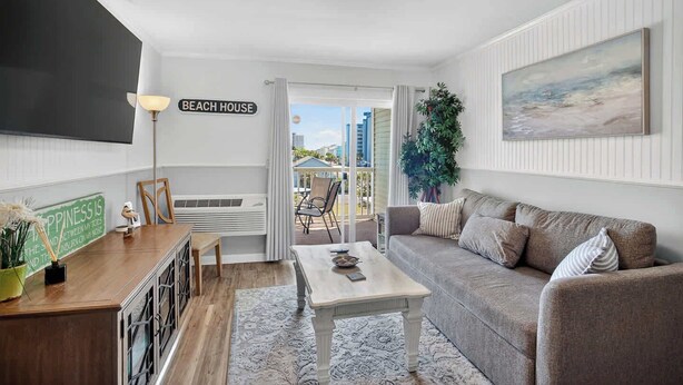 A modern living room with a sofa, coffee table, and a flat-screen TV. There's a sign on the wall that reads 'BEACH HOUSE' and a large window offering a view of the outdoors.