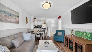 A living room with a sofa, a coffee table, a teal armchair, a TV, and a kitchen in the background.