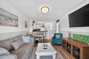 A living room with a sofa, a coffee table, a teal armchair, a TV, and a kitchen in the background. - North Myrtle Beach 2BR condo Steps from Beach (North Myrtle Beach)