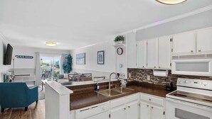 A modern kitchen with white cabinets, a microwave, and a stove. There is a sink with a faucet, a countertop, and a chair in the dining area. A clock and a plant are on the wall.
