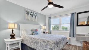A bedroom with a bed, two bedside tables with lamps, a ceiling fan, a window with curtains, and artwork on the wall.