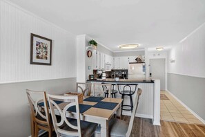 A modern kitchen with white cabinetry, a central island, and a dining area with a table and chairs. - North Myrtle Beach 2BR condo Steps from Beach (North Myrtle Beach)