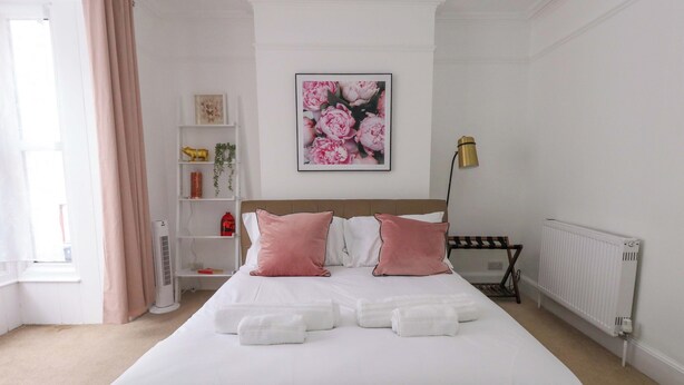Uma cama bem arrumada com almofadas brancas e rosa, uma pintura floral emoldurada acima e um radiador na parede.