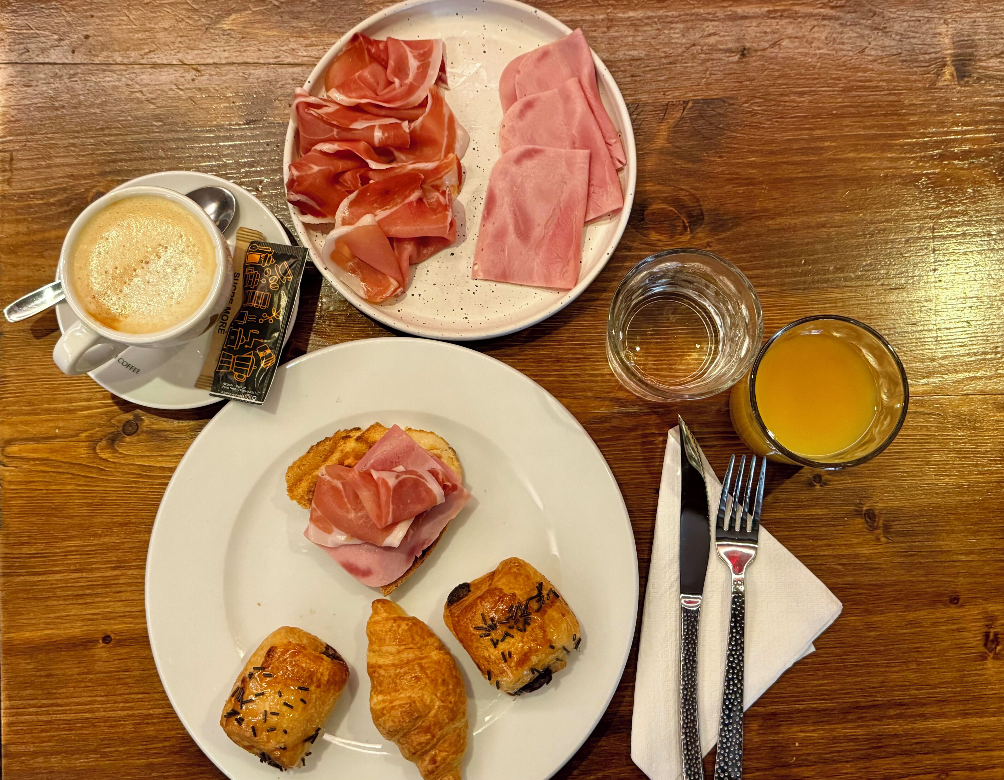 A breakfast setting with coffee, prosciutto, croissants, and juice on a wooden table.