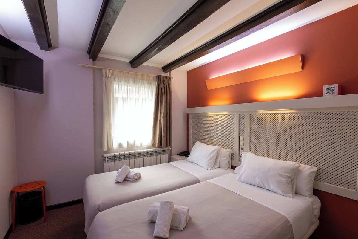 Habitación de hotel con dos camas, televisor, ventana con cortinas y una lámpara fijada a la pared.