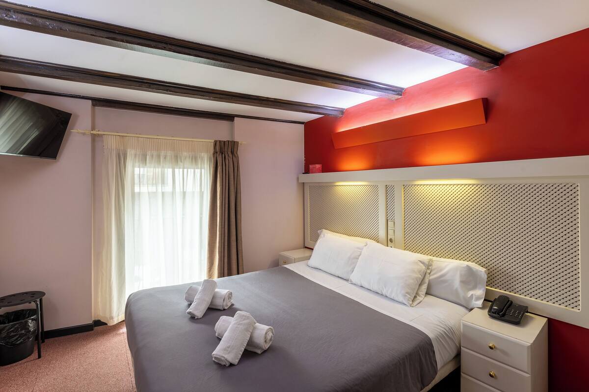 Habitación de hotel con cama, mesita de noche con teléfono, televisor montado en la pared y ventana con cortinas.