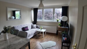 A living room with a grey sofa, a white coffee table, a small rug, a side table with a lamp, a mirror, and a window with curtains.