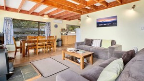 A cozy living room with a wooden dining table, grey sofa, and a view of the outdoors through large windows.