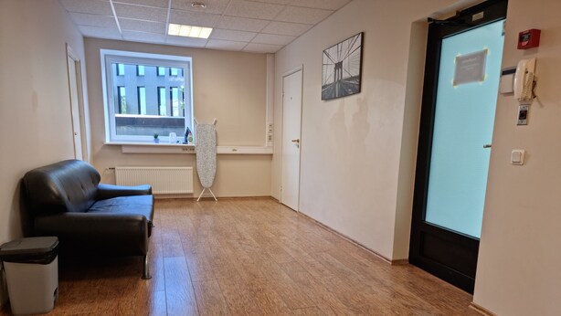 Une pièce avec un plancher en bois, un canapé en cuir noir, un radiateur, une fenêtre, une porte et un téléphone fixé au mur.