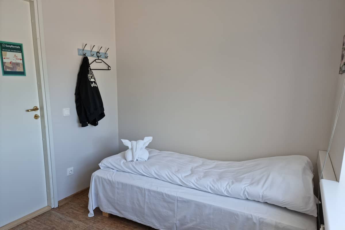 Een kleine, nette kamer met een enkel bed, een kapstok met een jas, en een deur met een bordje.
