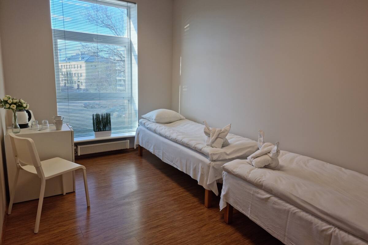 Een hotelkamer met twee enkele bedden, een witte stoel, een tafeltje met een bloevas en een raam met jaloezieën.