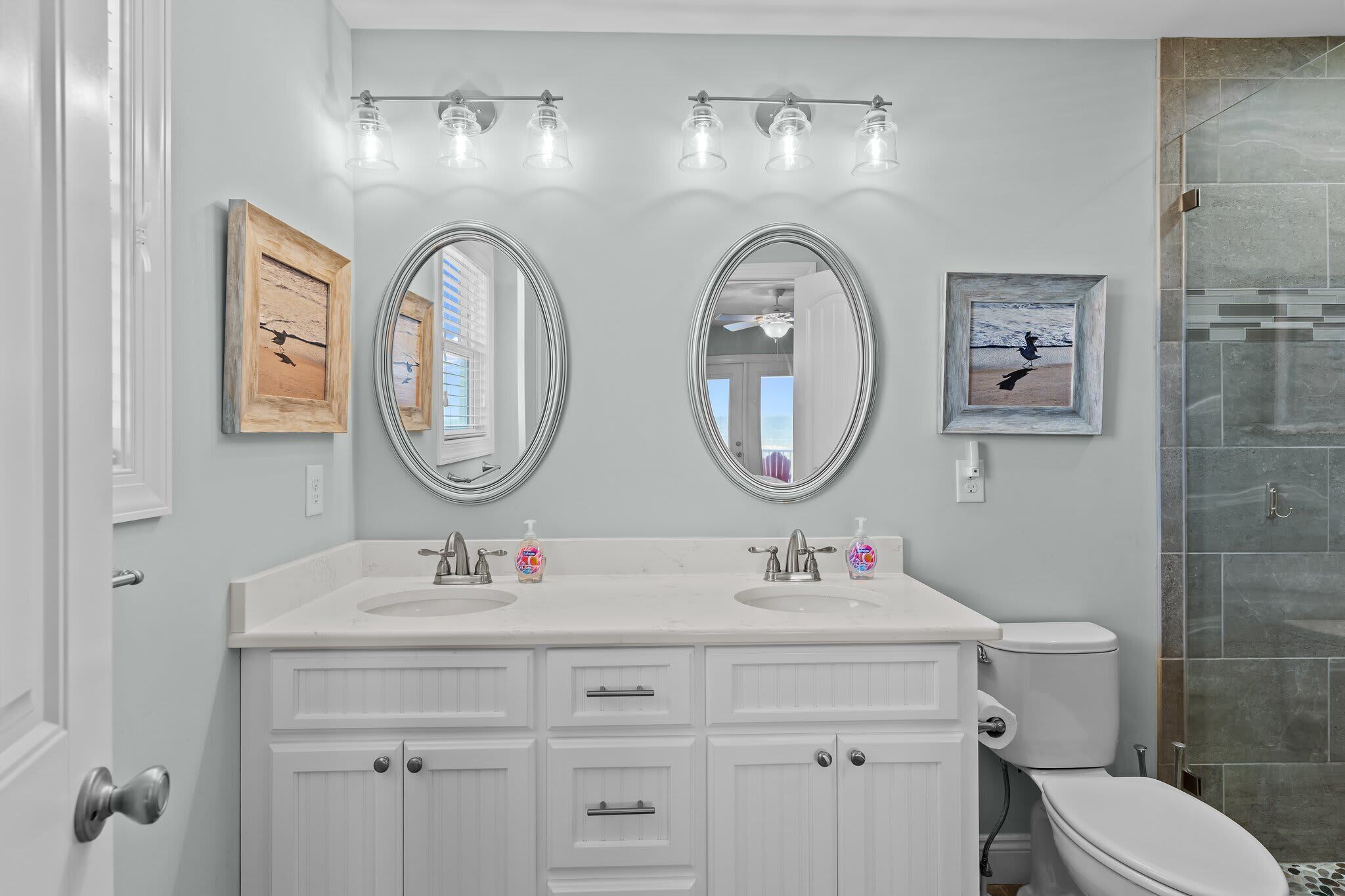 Een badkamer met twee wasbakken, een toilet en een douche. Er hangen afbeeldingen in lijsten aan de muur en een lamp boven de wasbakken.