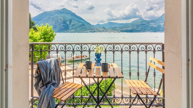 Un balcon avec vue sur un lac et des montagnes, des meubles en bois et un vase rempli de fleurs.