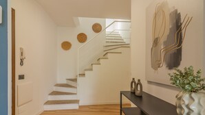 Un escalier avec des éléments en bois, un mur blanc orné de trois tentures tissées, et une console sombre sur laquelle sont posés un vase et deux bouteilles.