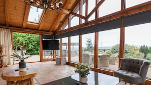 Una habitación amplia con un gran ventanal, una mesa negra, un sillón gris y un televisor sobre un trípode.