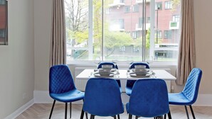 Un coin repas avec une table dressée pour quatre personnes, des chaises bleues et une vue sur un immeuble à travers de grandes fenêtres.