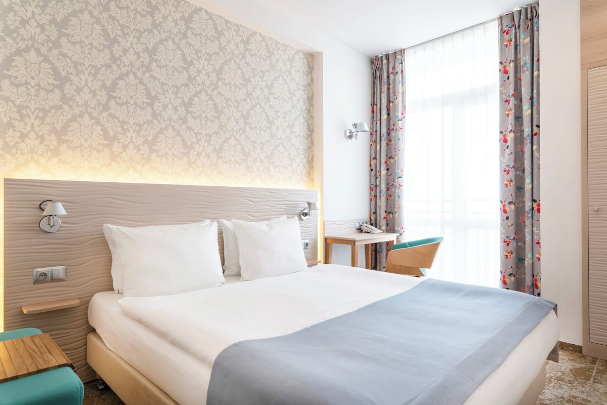 Una habitación de hotel con una cama grande, un escritorio, una silla y un ventanal con cortinas.