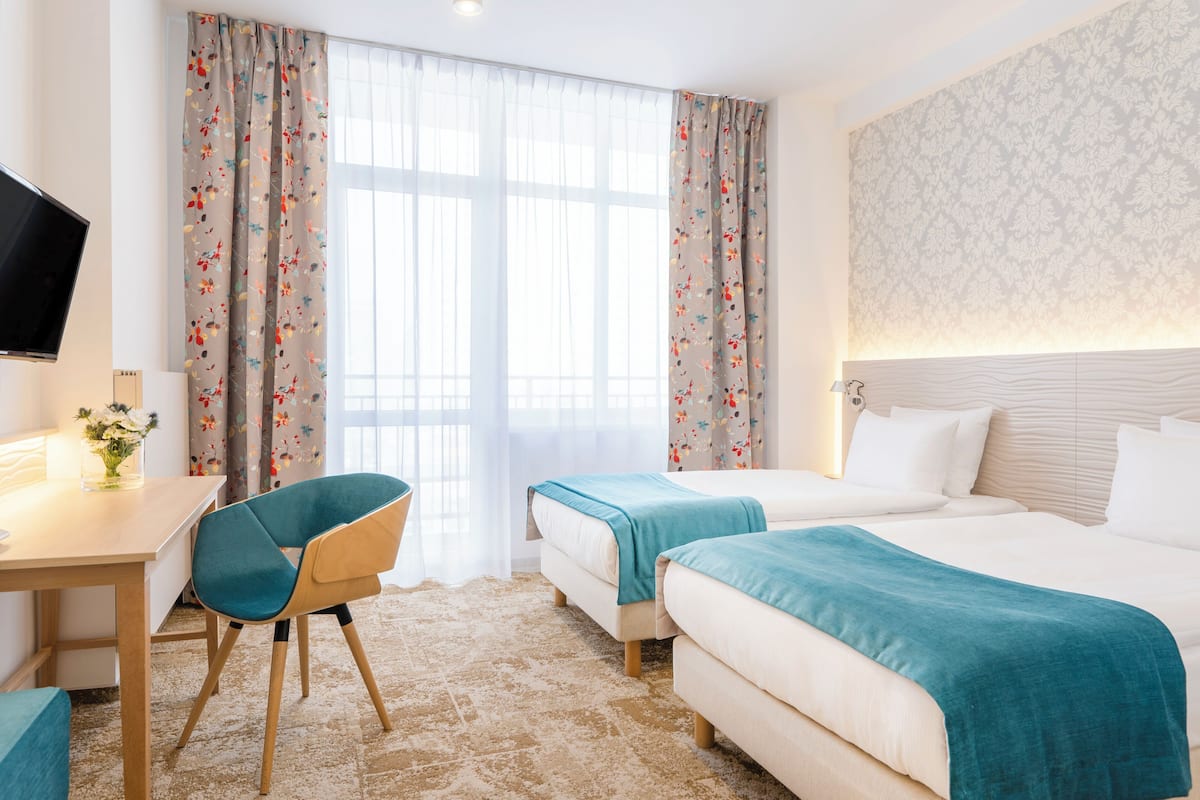 Una habitación de hotel con dos camas, un escritorio con silla, un televisor y una ventana con cortinas.