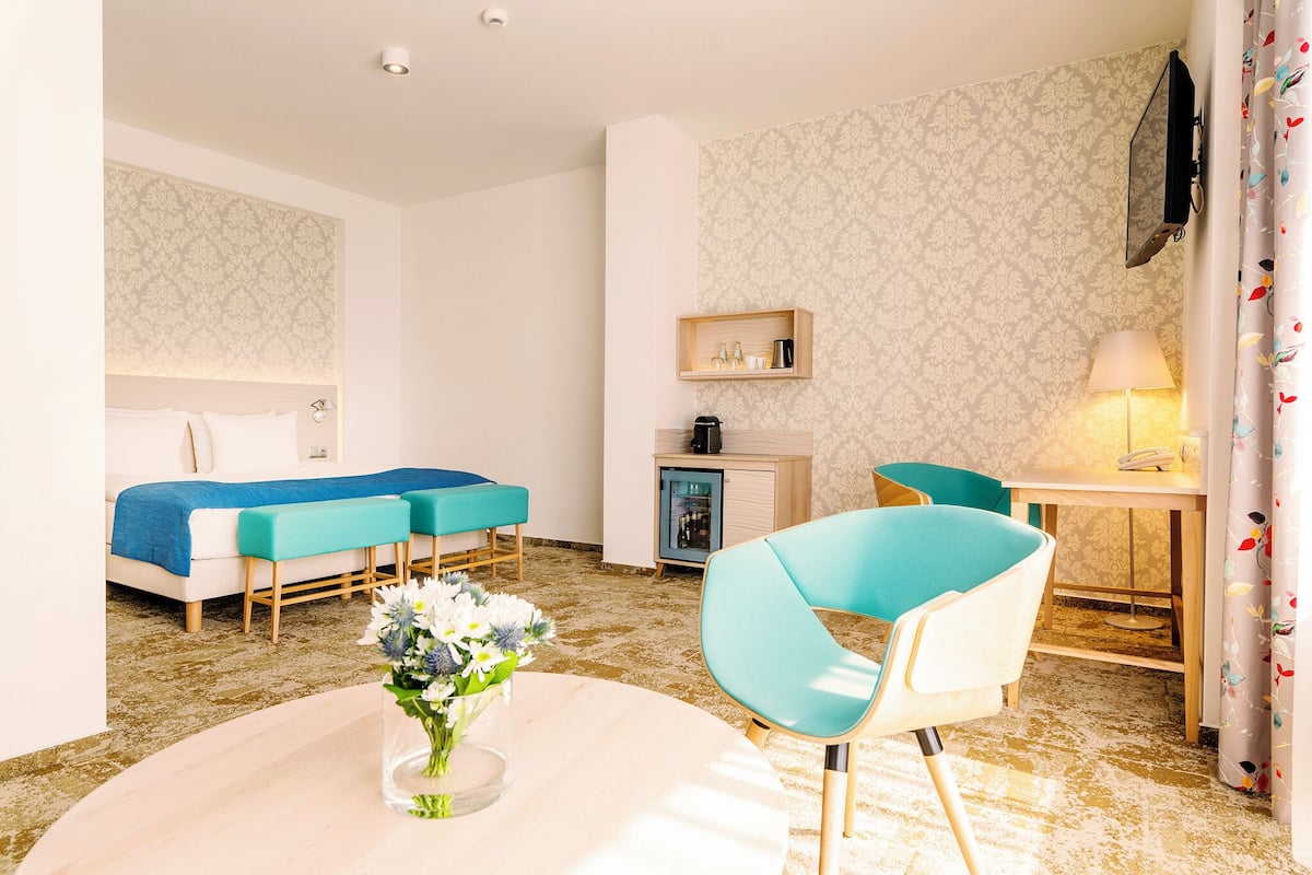 Habitación de hotel con cama, escritorio, silla, un mueble pequeño y un florero con flores.