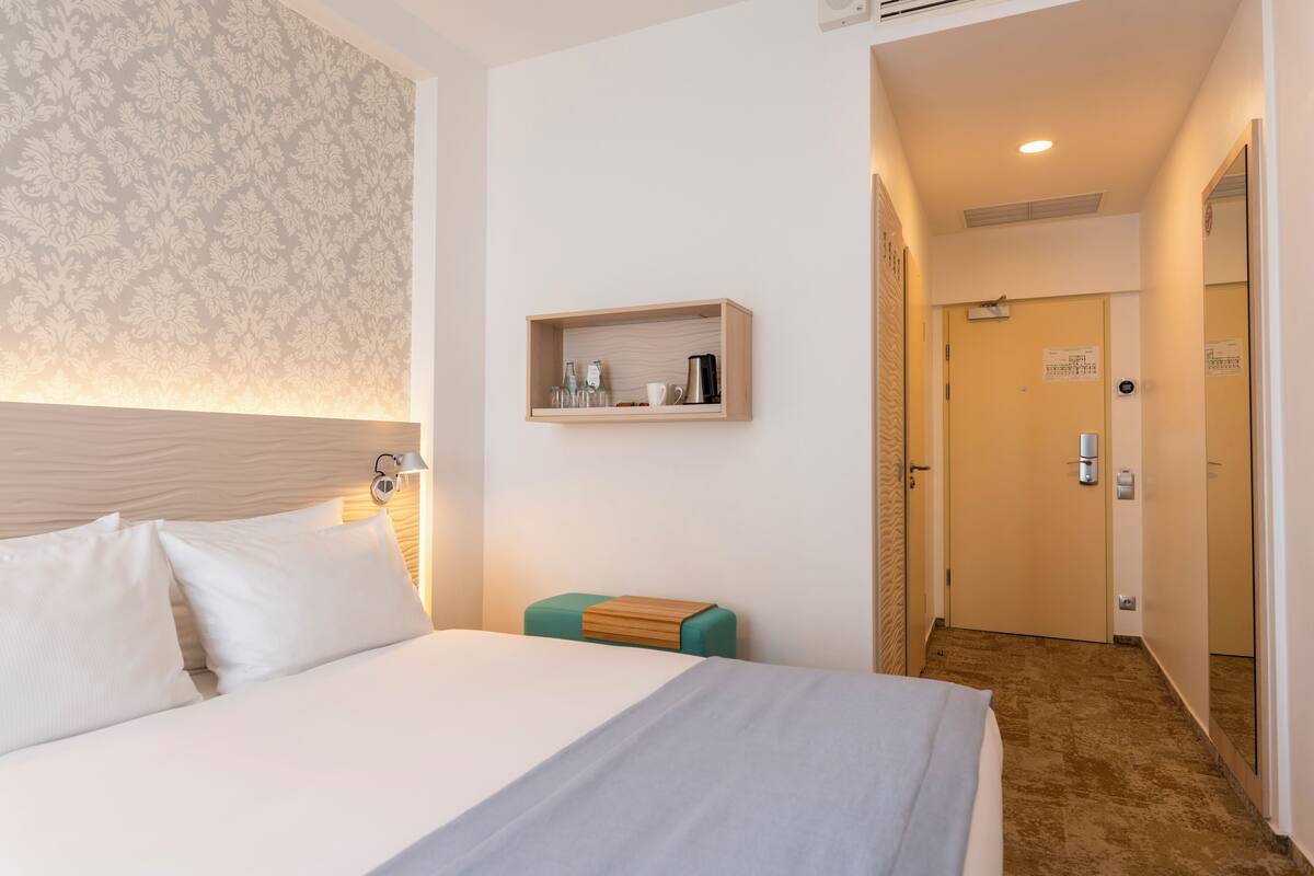Una habitación de hotel con una cama grande, una mesita de noche con lámpara, un pequeño estante de madera sobre la cama y una puerta que da a otra habitación.