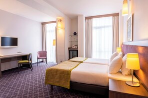 Et hotelværelse med to senge, et skrivebord, en stol, et fjernsyn og et badeværelse med et spejl og hylder.