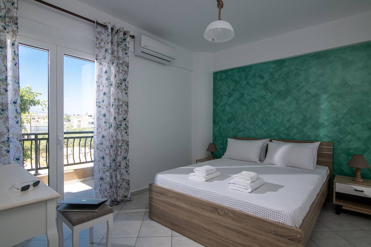 A bedroom with a bed, a desk, and a chair. There is a window with curtains and an air conditioning unit.