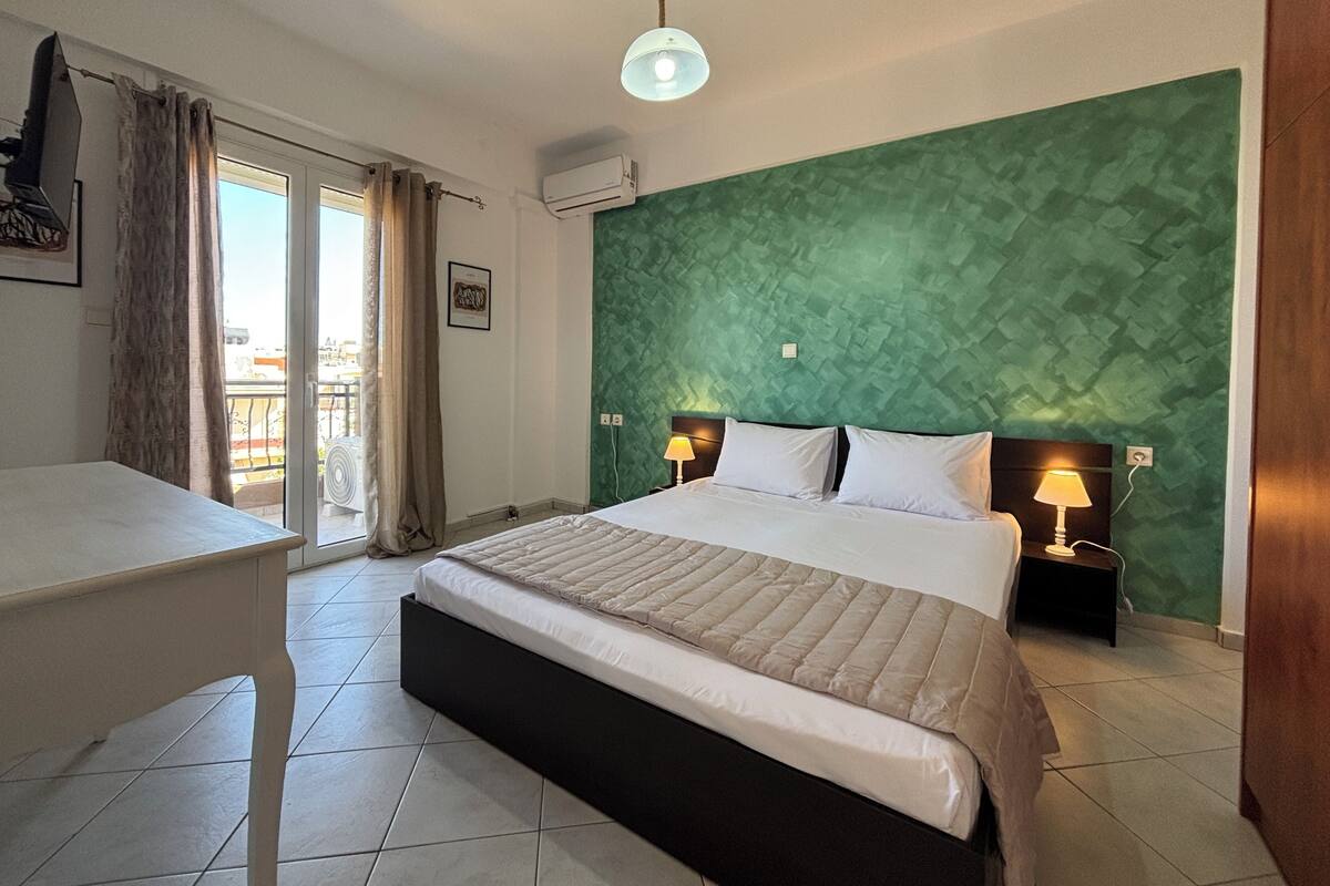A hotel room with a bed, bedside tables, a desk, a green textured wall, and a view through a sliding door.