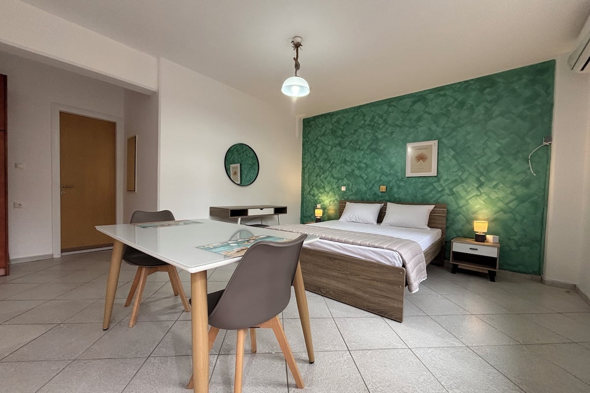 A modern bedroom with a green textured wall, a bed with white bedding, a bedside table with a lamp, and a dining area with a table and chairs.